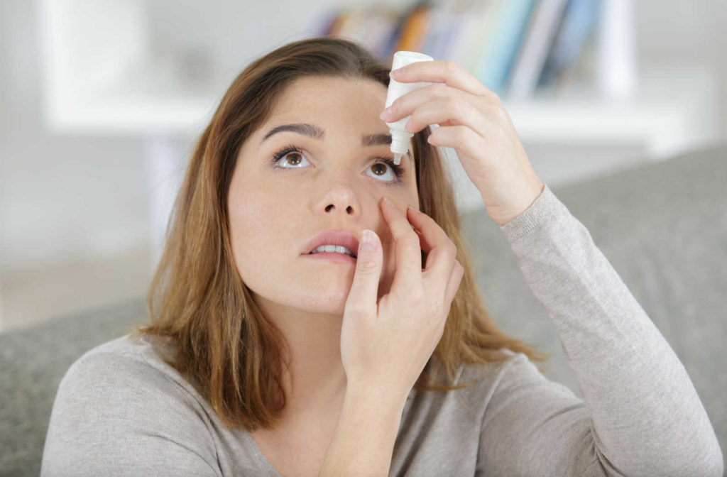 A woman holding a small bottle of eye drops in her left hand and putting them on her left eye