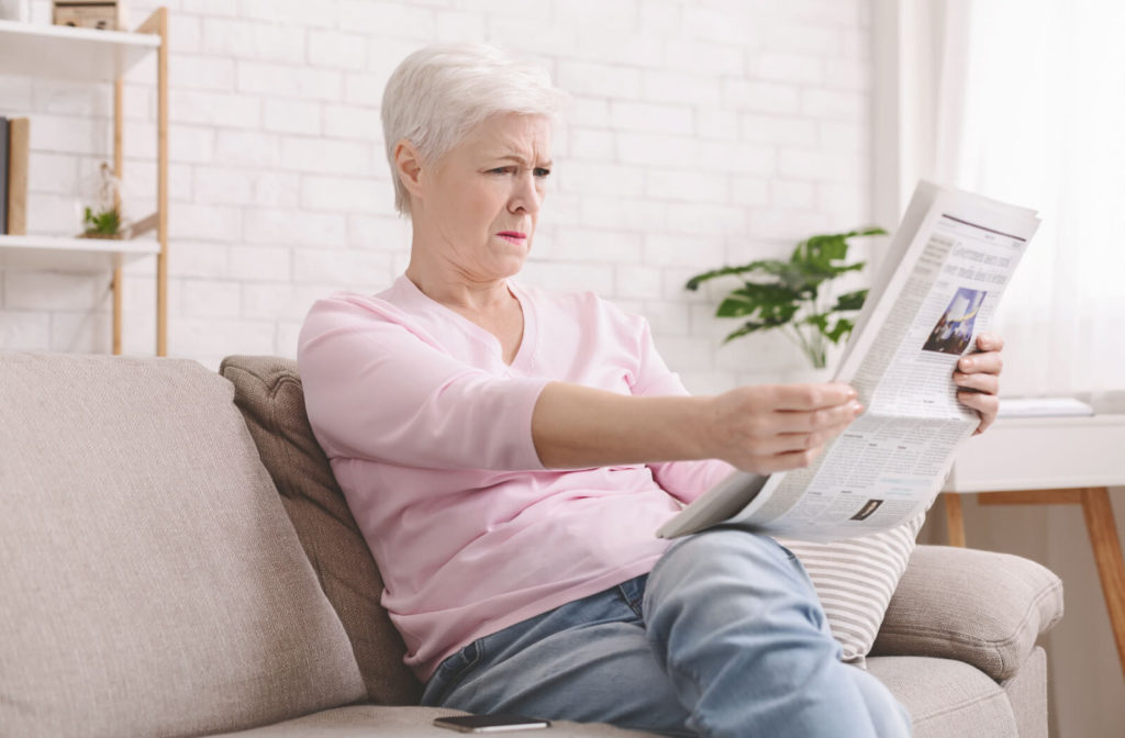 A senior woman squinting and holding a newspaper at arm's length because of vision challenges caused by presbyopia.