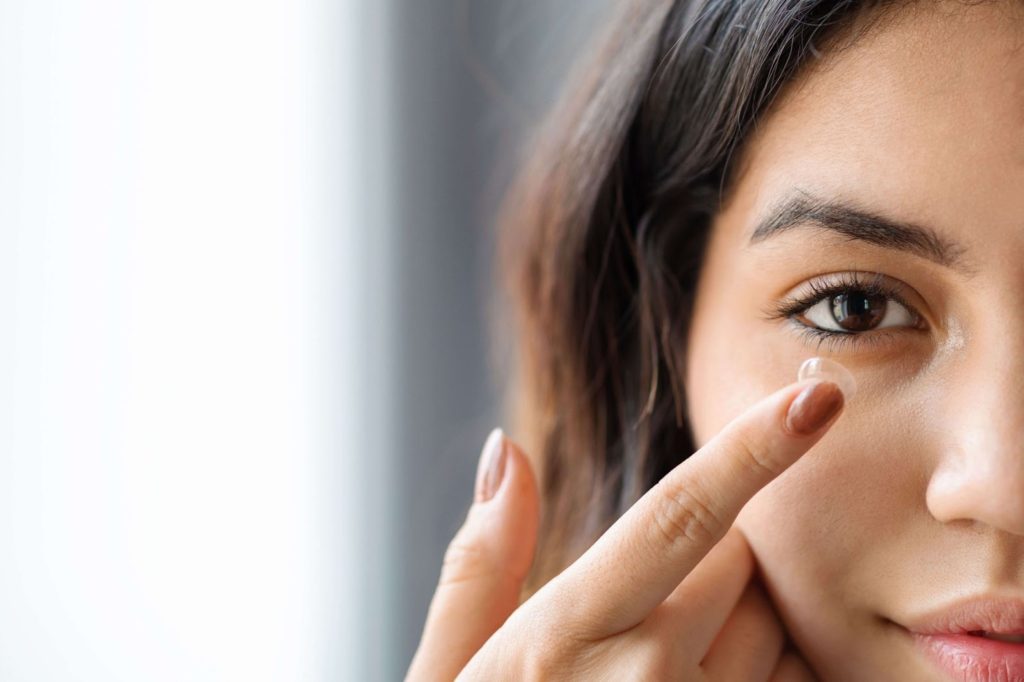 A close-up of the right side of a woman's face as she uses her index finger to place a contact lens on her eye.