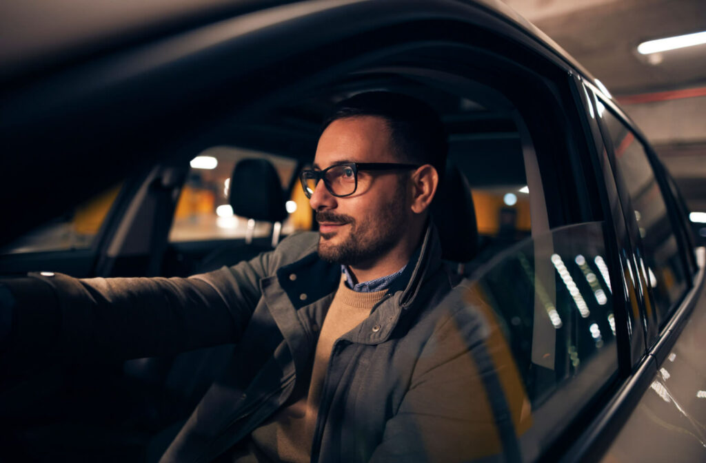 A young man with eyeglasses driving a car at night in a parking area.