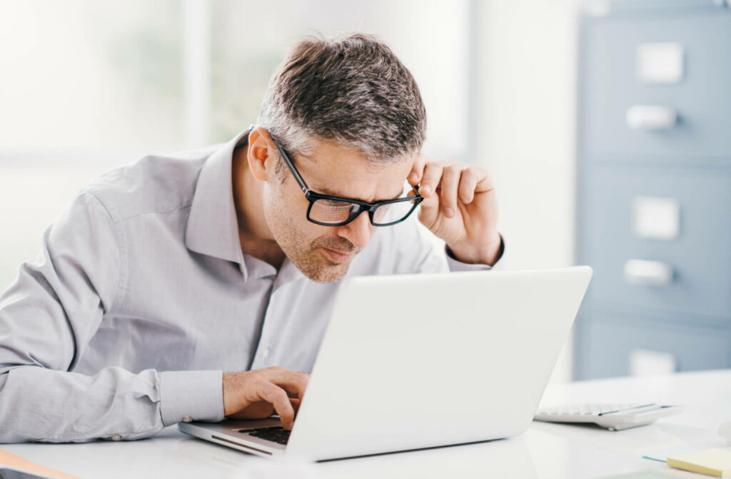 A man holding his eyeglasses with his left hand, squinting to see clearly on his laptop.