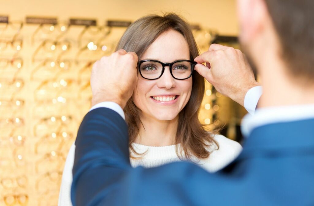 A young woman smiling and trying on glasses in a store while being assisted by an optician or optometrist.