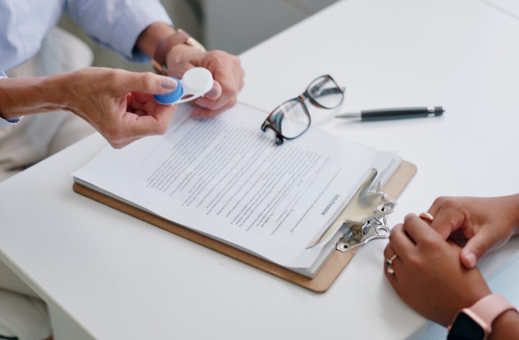 Looking down on a clipboard with eyeglasses sitting on it while an optometrist holds out a contact lens case.