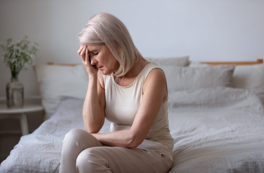 A senior sits on the edge of their bed with a hand on their head, suffering from headaches from high eye pressure