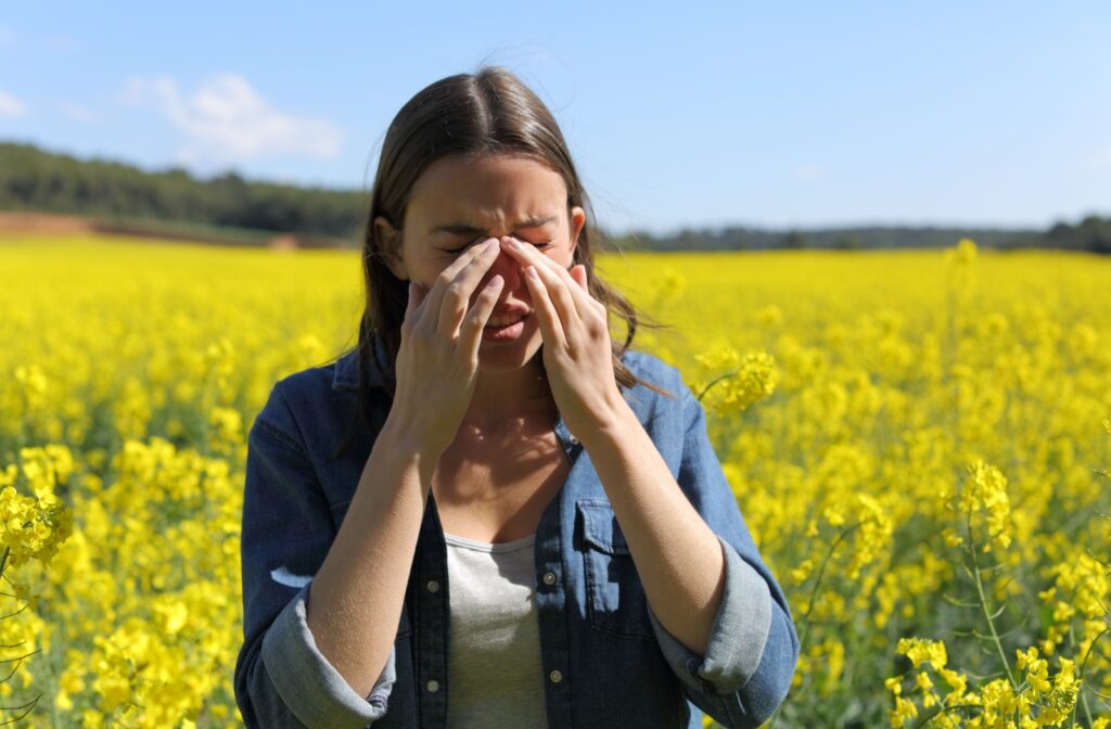 A young adult rubs at their eyes as they start itching while outdoors in a field of yellow flowers