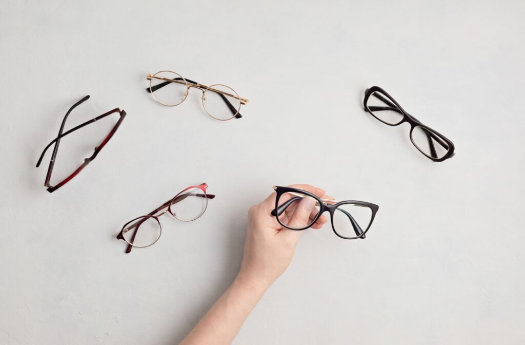 A variety of eyeglass styles sitting on a white desk top with a hand reaching out to grab a pair.