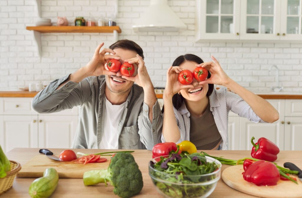 Two people holding up tomatoes over their eyes while standing in front of a table full of vegetables, symbolizing the importance of a healthy diet for eye health.